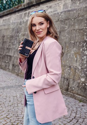 Black wallet with polka dots and tan accents held in a light pink blazer and denim jeans, against a textured stone background.