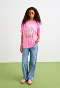 T-shirt rose surdimensionné avec un design graphique, associé à un jean large bleu clair et des baskets roses. Le mannequin porte des lunettes de soleil à monture marron.