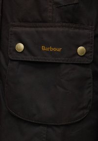 Poche de veste Barbour noire avec rabat sécurisé par deux boutons en laiton, logo Barbour brodé en jaune sur le rabat.