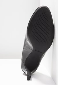 Chaussure à talon haut en cuir noir reposant sur la semelle contre un mur blanc d'angle, montrant le motif ondulé de la semelle et le logo "Clarks".