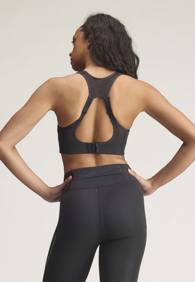 Kvinde med langt hår iført en sort racerback sports-bh og sorte højttaljede leggings, stående med hænderne på hofterne, ryggen vendt mod beskueren.