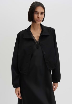 Frau mit kurzen dunklen Haaren, die eine schwarze, kurz geschnittene, geknöpfte Jacke über einem schwarzen Spitzenkleid vor einem einfarbigen hellen Hintergrund trägt.