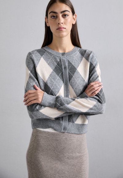 Tânără cu păr lung și închis la culoare poartă un cardigan argyle gri și crem și o fustă bej, stând cu brațele încrucișate pe un fundal simplu.