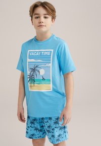 Ljusblå bomullströja med tryckt strandscen som har palmer. Matchas med blå shorts prydda med ett palmersmönster.