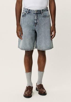 Hellblaue Jeansshorts mit lockerem Schnitt, mittlerer Bundhöhe und sichtbaren Nähten. Gestylt mit grauen Socken und braunen Schnürschuhen.