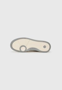 Sola de ténis em borracha bege e cinza, com um padrão em zigue-zague, detalhes circulares e o nome da marca "Lacoste" em relevo perto do calcanhar.