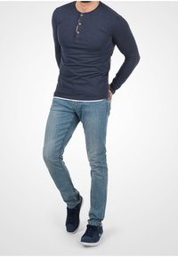 Hombre vistiendo una camiseta henley de manga larga azul marino, jeans de color azul claro y zapatillas azul marino, de pie con las manos detrás de la espalda sobre un fondo blanco.