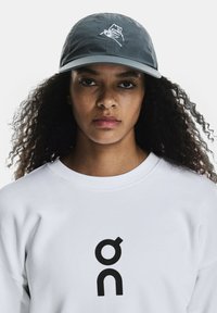 Căciulă gri cu un logo frontal, având un design cusut. Purtaată cu un hanorac alb care afișează un logo negru proeminent al mărcii.