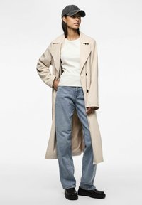 Frau in beigem Trenchcoat, weißem Oberteil, hellblauen Jeans, schwarzen Stiefeln und dunkelgrauem Baseballcap, steht mit einer Hand in der Tasche.