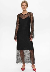 Černé krajkové šaty s dlouhými průhlednými rukávy, pod nimiž se nachází jednobarevný černý podšívkový model. Lem je asymetrický, odhalující krajkové detaily.