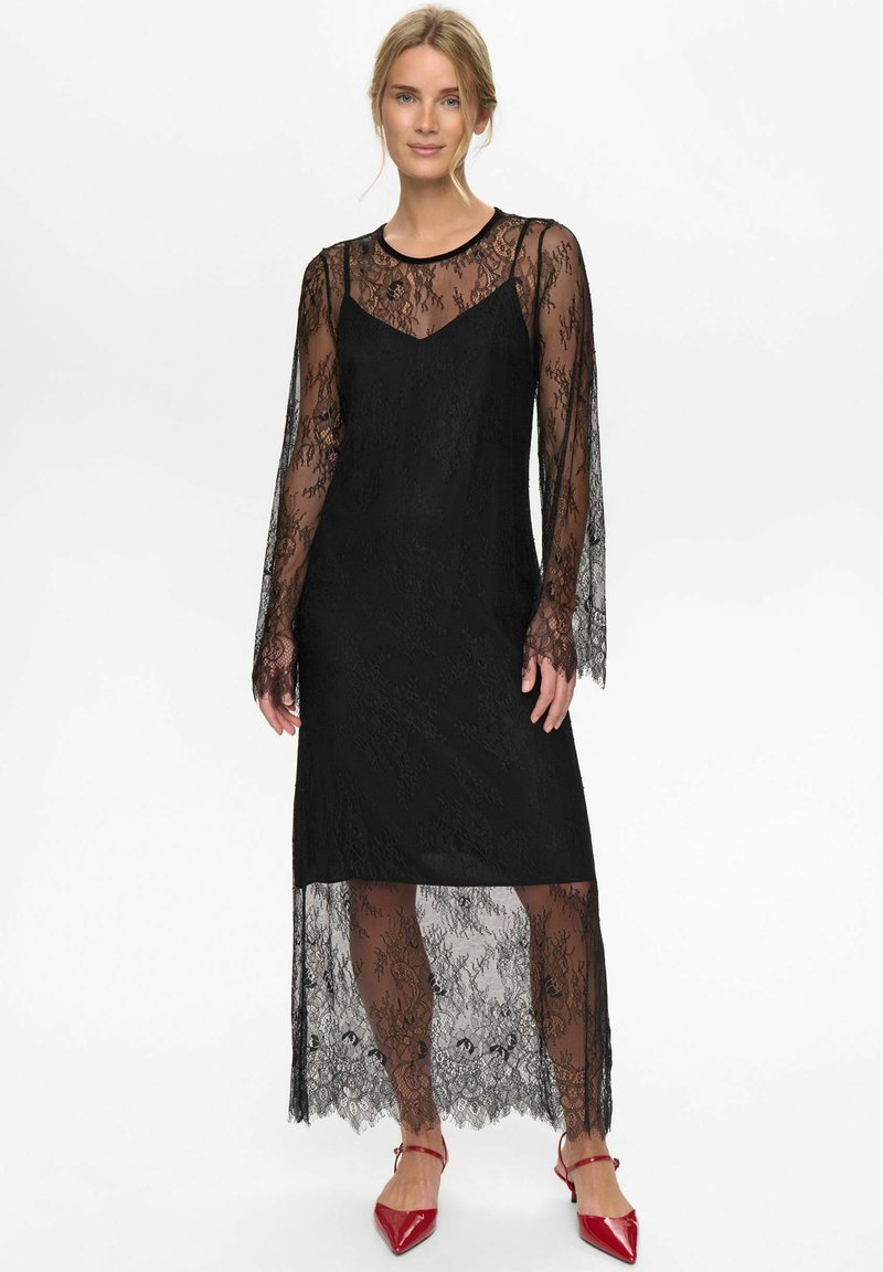 Černé krajkové šaty s dlouhými průhlednými rukávy, pod nimiž se nachází jednobarevný černý podšívkový model. Lem je asymetrický, odhalující krajkové detaily.