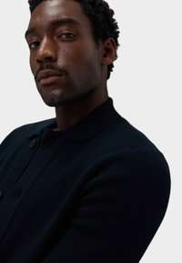 Navyblauer Strickpullover mit Kragen, der über einen Knopf und einen strukturierten Stoff verfügt. Nahaufnahme des Kleidungsstücks und der Schulter.