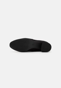 Zapato negro con una textura de ante suave, punta redondeada y tacón apilado. La suela presenta un agarre con patrón para mayor tracción.