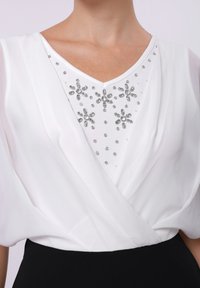 White blouse with a V-neck, featuring a jeweled floral design on the bodice. The fabric is lightweight and drapes softly around the neckline.