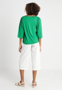 Groene top met korte mouwen en geplooide details op de schouders, gecombineerd met witte wijde culottes. Model draagt beige sandalen.