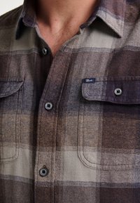 Camicia in flanella a quadri nei toni terrosi del marrone e del beige. Caratterizzata da chiusura con bottoni, due tasche frontali e un piccolo etichetta del marchio sul petto.