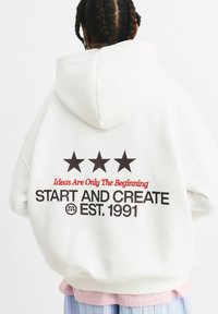 Witte oversized hoodie met drie zwarte sterren. De tekst op de achterkant luidt "Ideeën zijn slechts het begin" en "START EN CREËER EST. 1991" in vette zwart- en roodkleurige letters.