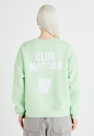 Piparmētru zaļš džemperis ar pārspīlētu piegriezumu, apaļu kakla izgriezumu un baltu grafiku ar tekstu "CLUB MATCHA" un krūzes ilustrāciju uz muguras.