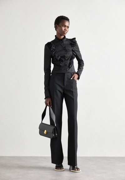 Čierna volániková blúzka s gombíkovým zapínaním, skombinovaná s priliehavými čiernymi nohavicami. Doplnky zahŕňajú pevný čierny kabelku so zlatým hardvérom.