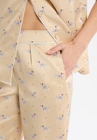 Beżowy jedwabny strój składający się z krótkiego topu i spodni, ozdobiony małymi wzorami czarnych i białych psów oraz motywami diamentów w kontrastowym kolorze.