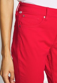 Pantalons en mélange de coton rouge avec une texture lisse, dotés de poches frontales, d'une fermeture éclair et d'un bouton, et de détails de couture subtils.