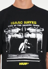 Sort bomulds t-shirt med et grafiktryk af Isaac Hayes, der optræder, med gul tekst, der siger "ISAAC HAYES LIVE AT THE SAHARA TAHOE."