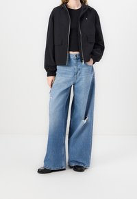 Czarna kurtka krótka z kapturem i przednimi kieszeniami, połączona z jasnoniebieskimi spodniami o szerokich nogawkach z dziurami. Model ma na sobie czarne buty.