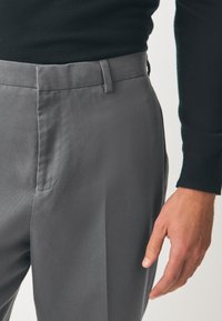 Pantalon gris sur mesure avec une texture lisse, des lignes épurées et des poches latérales, associé à un haut noir à manches longues ajusté.