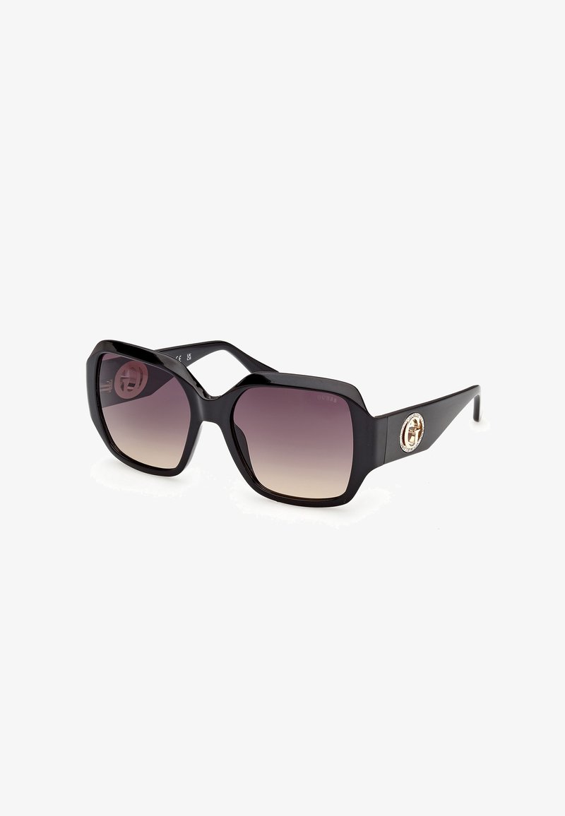 Lunettes de soleil carrées surdimensionnées noires avec des verres marron dégradés et un logo circulaire doré sur la branche.