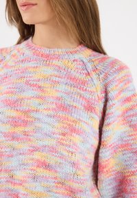 Femme portant un pull en tricot pastel multicolore aux teintes rose, violet, jaune et bleu, montrant le haut du torse et le cou.