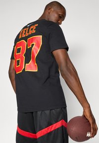 Sort t-shirt med r�ød og gul grafik "KELCE 87" på ryggen. Modellen holder en brun fodbold og har sorte shorts med en rød stribe.
