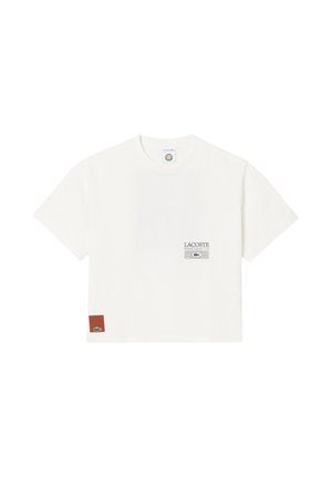 Camiseta blanca de manga corta con cuello redondo, pequeño logo del Lacoste Roland Garros Club en el pecho y parche marrón con el logo del cocodrilo verde cerca del dobladillo.