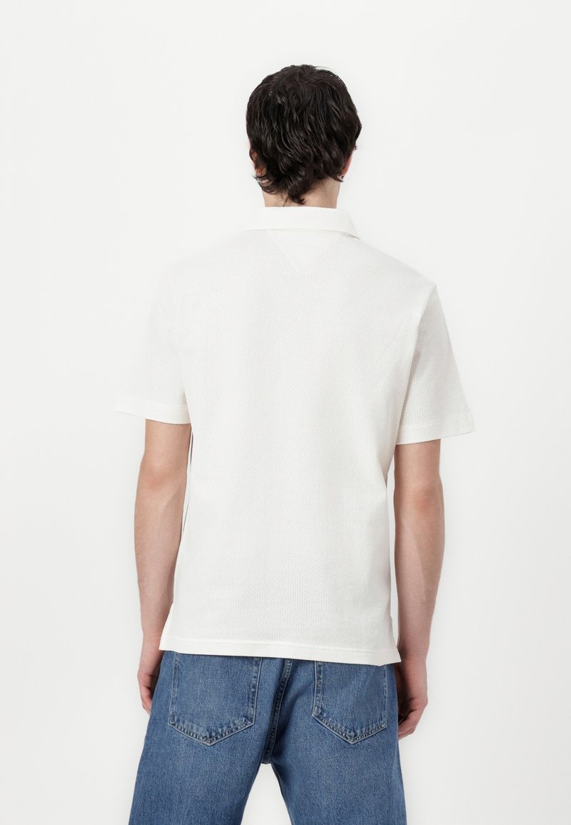 Homme aux cheveux foncés portant une chemise blanche à manches courtes et un jean bleu, debout dos à un fond blanc uni.