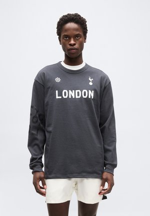 Person iført en mørkegrå langærmet trøje med teksten "LONDON" og hvide shorts, stående mod en ensfarvet lys baggrund.