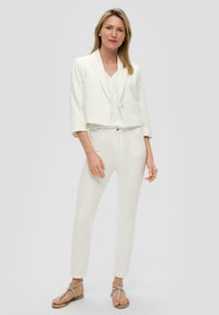Witte cropped blazer over een wit patroontop en slim-fit witte broek. Model draagt zilveren sandalen met bandjes. Eenzelvig grijze achtergrond.