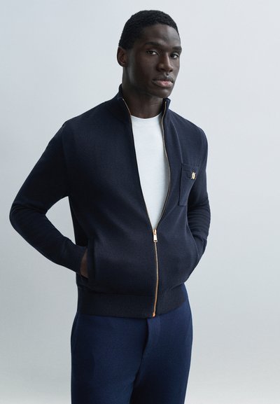 Homme portant une veste zippée bleu marine sur une chemise blanche, debout les mains dans les poches, devant un fond gris clair uni.