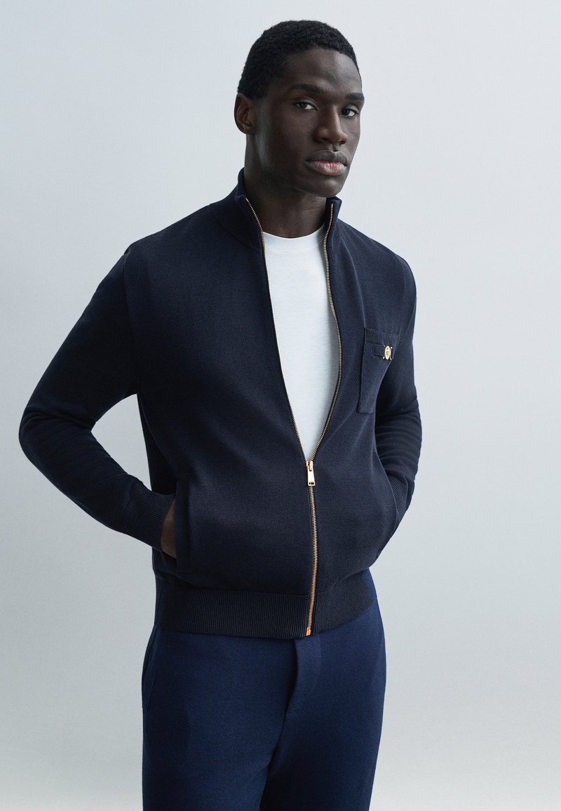 Vyras, vilkintis tamsiai mėlyną užtrauktuku užsegamą striukę ant baltos marškinėlių, stovintis rankomis kišenėse priešais paprastą šviesiai pilką foną.