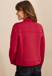 Röd jacka med en strukturerad design, som har en platt krage, sydda detaljer och ribbade ärmslut. Buren över mörka jeans, slät textur.