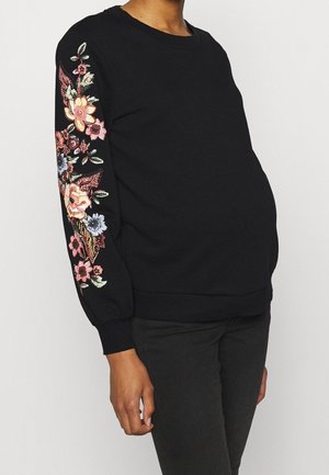 Persoon draagt een zwart longsleeve top met kleurrijke bloemenborduursels op de mouwen en een zwarte broek, staand tegen een effen achtergrond.