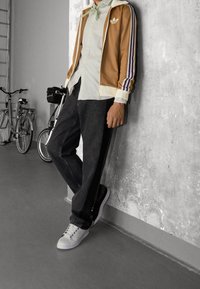 Brun idrottsjacka med vita och lila ränder, vit skjorta, mörka jeans och vita sneakers; står mot en betongvägg med cyklar i närheten.