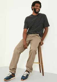 Black and navy striped short-sleeve shirt, beige pants, blue athletic shoes with tan accents, seated on a wooden stool against a light background.
