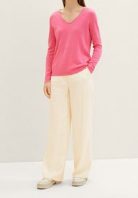 Rosa V-Ausschnitt Pullover mit langen Ärmeln, kombiniert mit cremefarbenen Hosen mit weitem Bein, die eine glatte Textur und eine entspannte Passform aufweisen. Leichte Schuhe vervollständigen den Look.