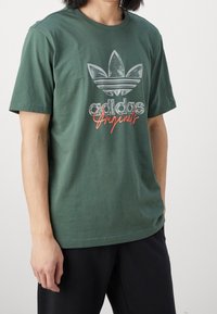 Zelené bavlněné tričko s krátkým rukávem a logem Adidas Originals v šedé a oranžové barvě. Disponuje kulatým výstřihem a volným střihem.