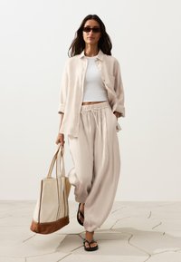 Femme marchant en pantalon ample beige, chemise ouverte assortie, crop top blanc, sandales noires, lunettes de soleil, portant un grand sac fourre-tout beige et marron.