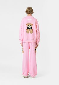 Conjunto de sudadera y pantalones rosa con un diseño de oso de peluche en la parte posterior, con un acabado texturizado y detalles en negro en el atuendo del oso.