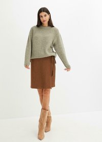 Maglione verde chiaro in peluche con spalle scese, abbinato a una gonna marrone con fiocco laterale. Modella che indossa stivali color crema al ginocchio.