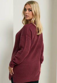 Übergroßer burgunderfarbener Strickpullover mit strukturiertem Muster, langen Ärmeln, seitlichen Schlitzen und einer lockeren Passform, getragen über schwarzen Hosen.