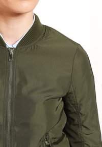 Bomber bunda v olivově zelené barvě má žebrovaný límec, zipové zapínání a hladkou texturu s předními kapsami a šitými detaily.