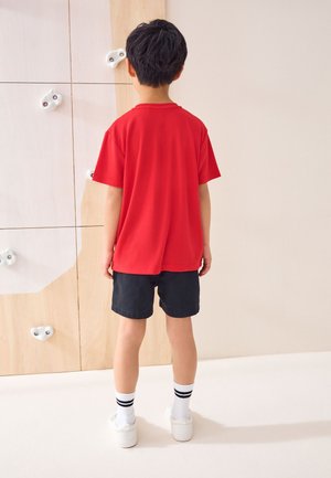 Piros ingben és fekete rövidnadrágban álló fiú, aki egy fehér fogantyúkkal ellátott beltéri mászófal felé néz.