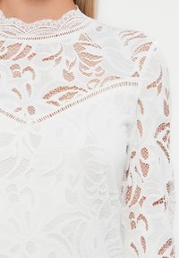 Top in pizzo bianco con motivi floreali, collo alto con bordo scallop e maniche lunghe con dettagli semitrasparenti.